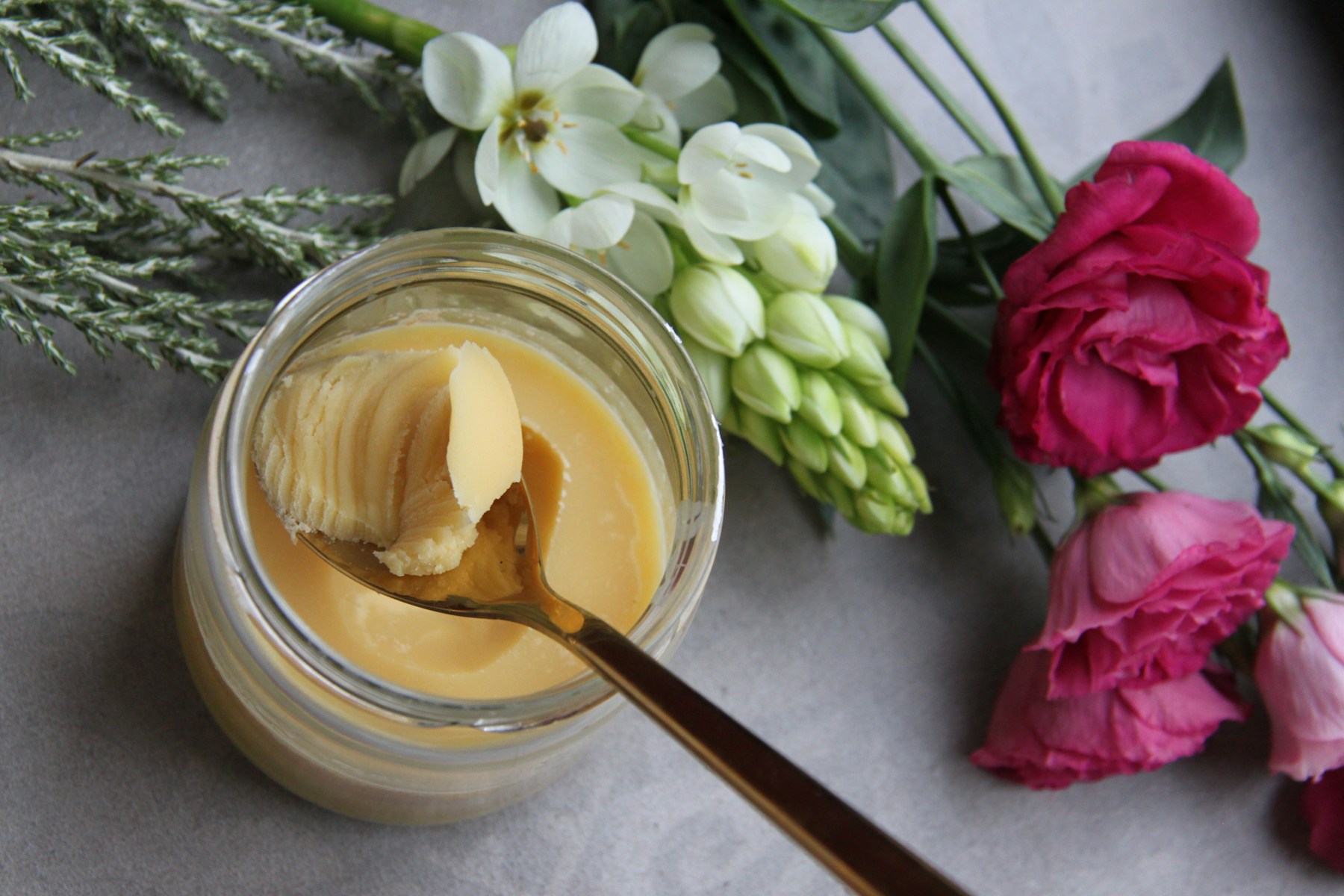 A glass jar of pale butter with a gold spoon, beside white and pink flowers