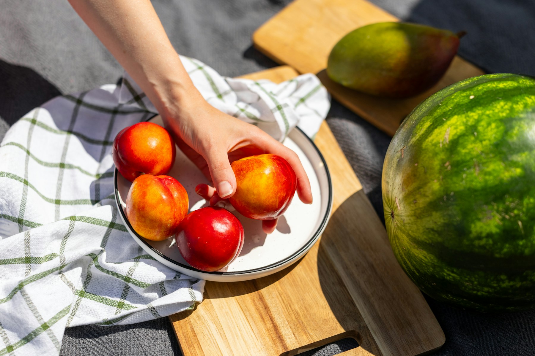 A hand picking nectarines from a bowl on a picnic cloth, with watermelon and mango nearby