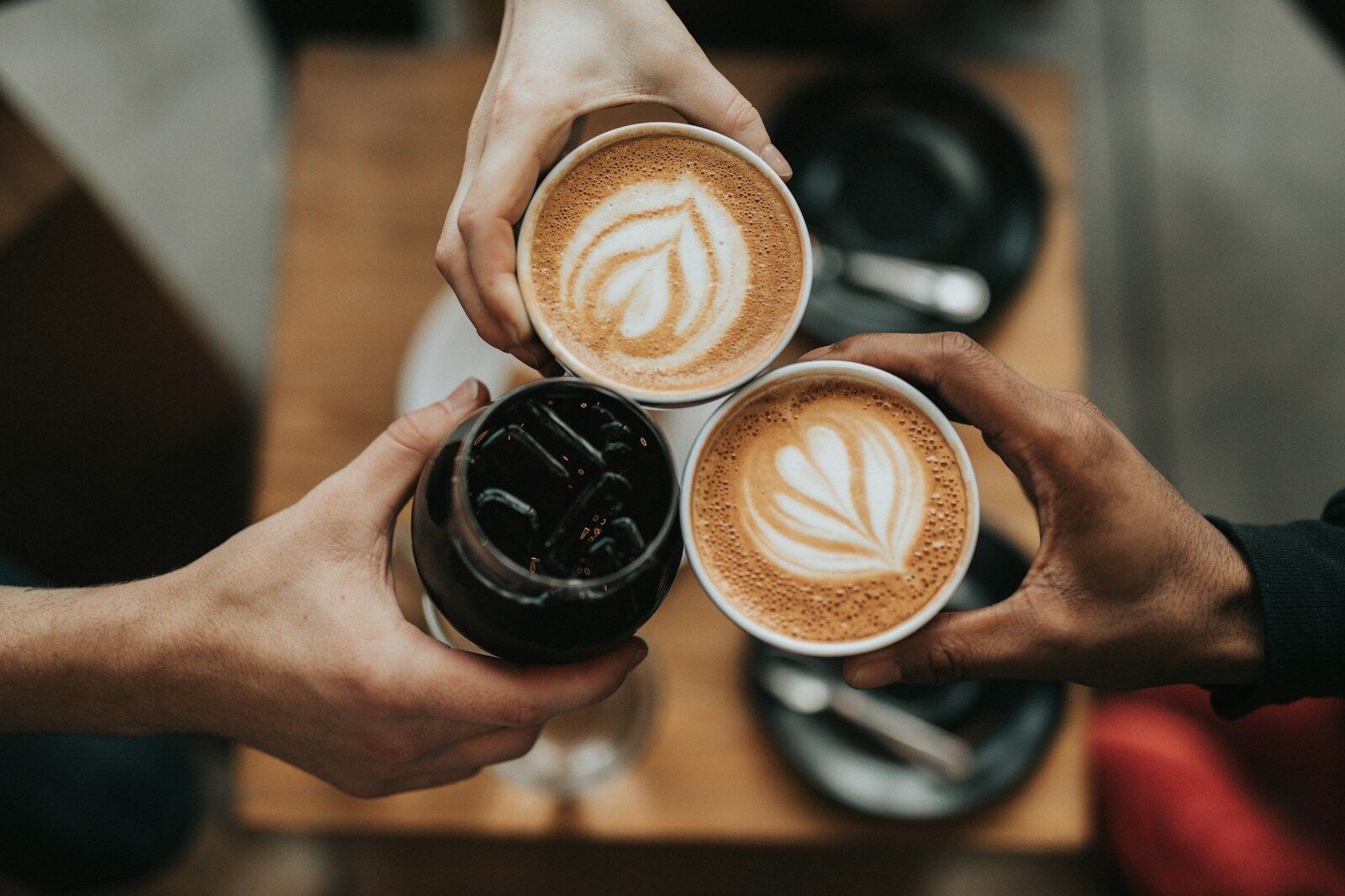 Hands raising two lattes with leaf art and an iced coffee over a wooden table
